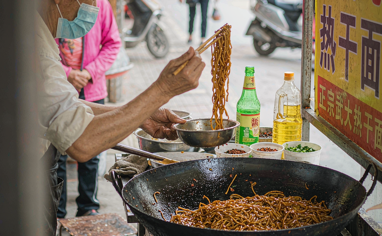 A vendor stirring sesame-paste hot dry noodles in a steel bowl at a Wuhan street breakfast stall