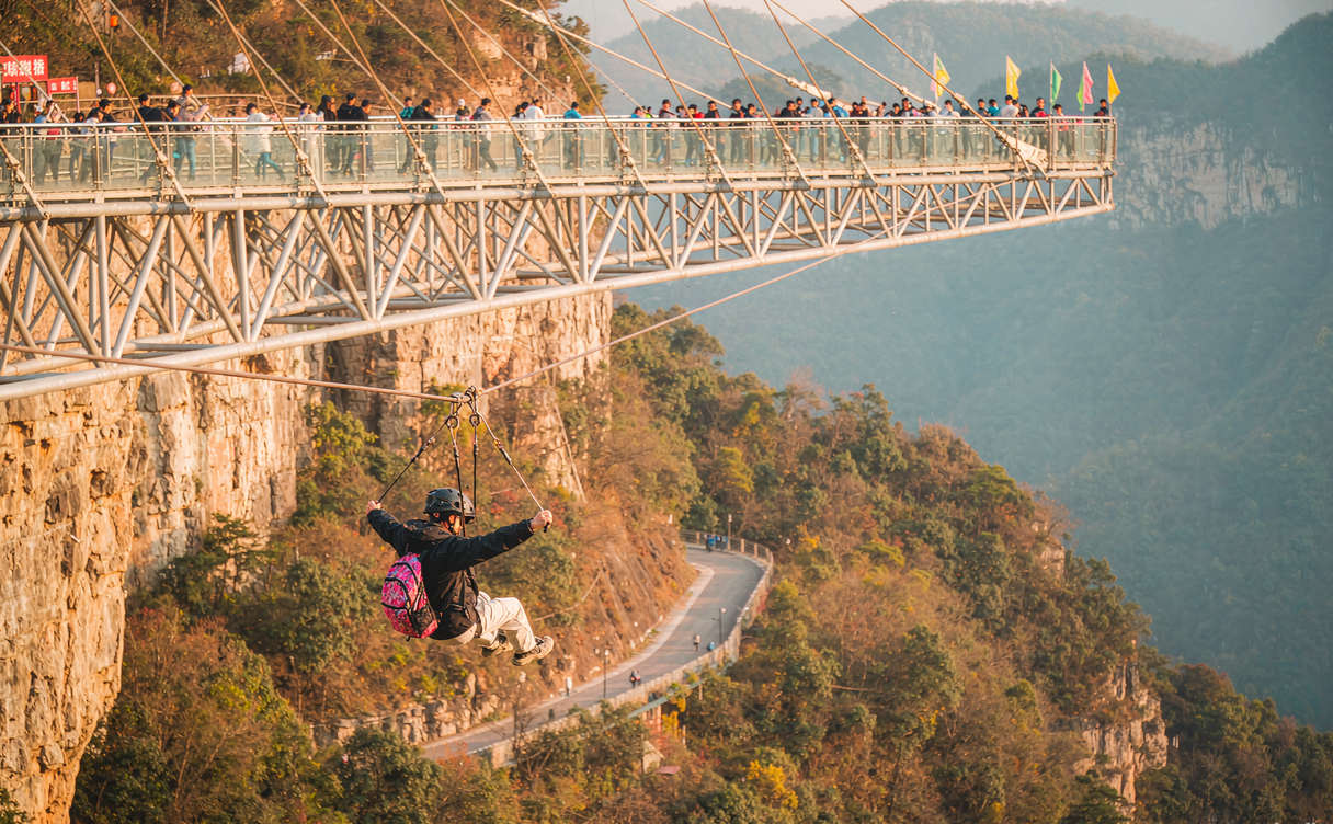 A zip line rider flying across the Zhangjiajie Grand Canyon with the glass bridge visible beside the cliffs in Hunan, China