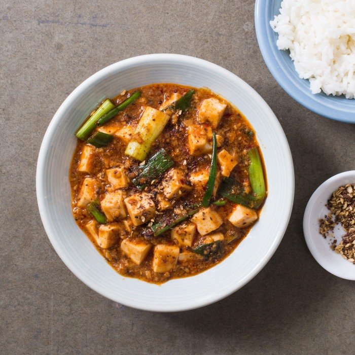 A bowl of Chengdu-style mapo tofu with tofu cubes in glossy red chili oil, scallions, served beside steamed rice and a dish of ground Sichuan peppercorn