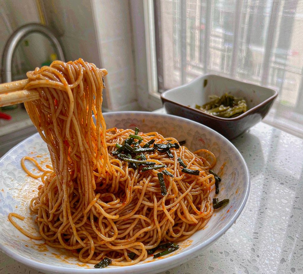 Chopsticks lifting scallion oil noodles from a bowl, with fried scallions and a side dish of pickled greens beside it