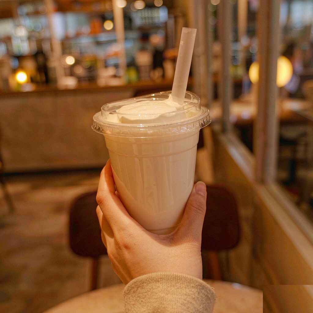 A hand holding a clear cup of milk tea topped with a thick layer of white cheese foam, in a softly lit cafe